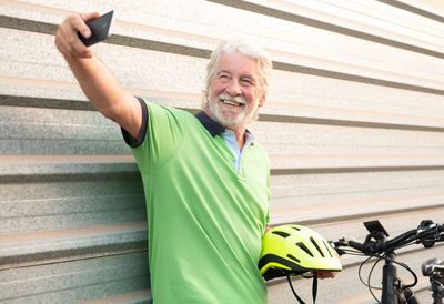 Smiling senior man taking selfie while holding cycling helmet against shutter