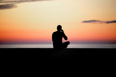 Silhouette man sitting at beach against sky during sunset
