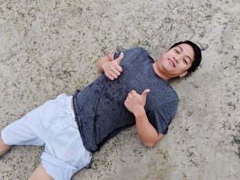 High angle portrait of man gesturing thumbs up sign while lying on sand