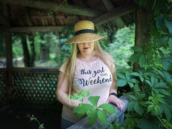 Portrait of young blonde woman wearing a hat standing in a wooden gazebo covered in green leaves