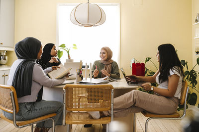 Smiling young woman studying with friends sitting at table in kitchen