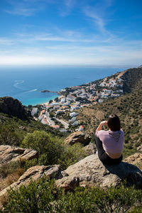 Rear view of woman sitting on rock by sea against sky