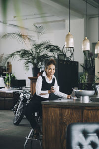 Portrait of smiling businesswoman holding coffee mug while sitting at kitchen island in creative office