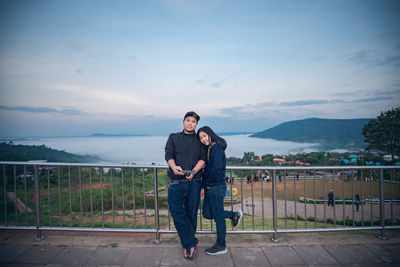 Full length portrait of couple standing against railing and sky