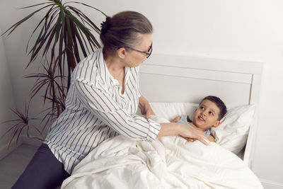 Grandmother in glasses and a white shirt reads a book to her grandson lying on the bed 