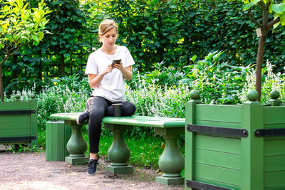 Young pretty blonde sits on bench in park with  and holds smartphone in her hands.