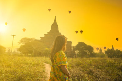 Rear view of woman with balloons at field against sky during sunset