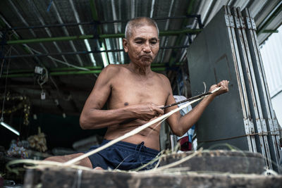 Shirtless senior man cutting plant on seat at market