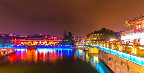 Illuminated bridge over river by buildings against sky at night