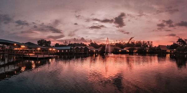 Illuminated buildings by river against sky at sunset