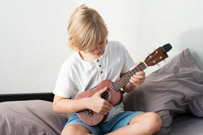 Young boy tuning ukulele at home. blond haired boy sitting on couch playing acoustic guitar.