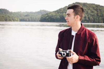 Young man looking away while holding old camera against lake and mountain