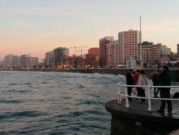 People on sea by buildings against sky during sunset