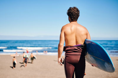 Back view of unrecognizable male athlete with surfboard admiring waving sea in sunny cloudless day