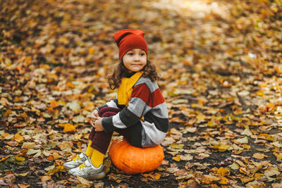Happy cute little girl child in warm bright clothes sitting on a pumpkin in the autumn forest