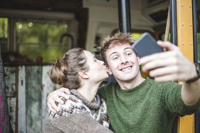 Smiling man taking selfie from mobile phone while woman kissing him during camping