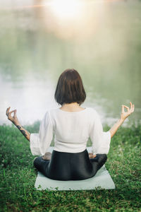 Young woman sitting in lotus position and practicing meditation near water in the nature