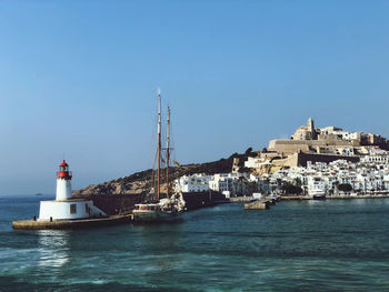 Lighthouse by sea against buildings against clear blue sky
