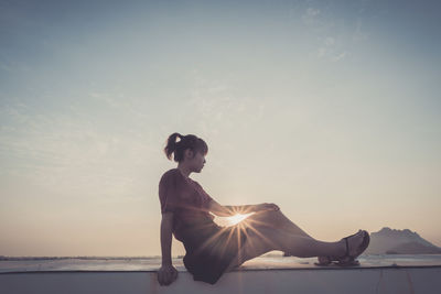 Woman sitting on retaining wall against sky during sunset