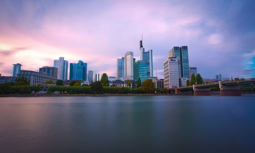 Buildings by river against sky during sunset