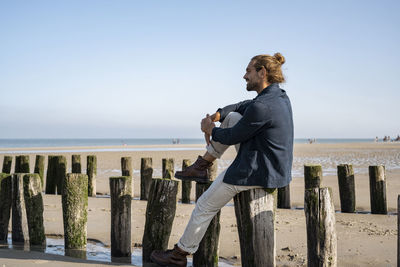 Side view of man on wooden post by sea against clear sky