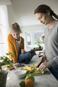 Girl looking at mother cutting fruits on kitchen counter at home