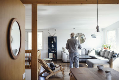 Rear view of senior man standing in living room at home on sunny day