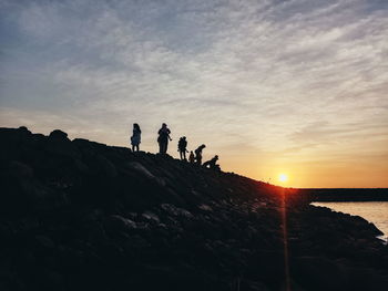 People on rocks by sea against sky during sunset