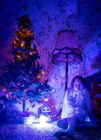 Girl sitting by illuminated christmas tree at night