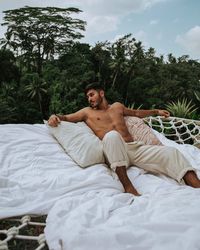 Shirtless young man relaxing on bed against trees in forest