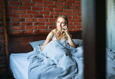 Adult woman forty years blonde in pajamas using mobile phone on bed in loft room at home