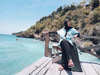 Portrait of young woman sitting on wood by sea against sky