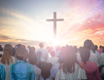 Digital composite image of crowd and cross against cloudy sky during sunset