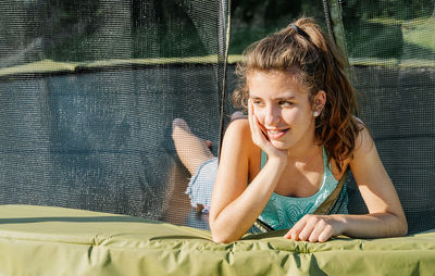 Horizontal portrait of a curly brunette teenager lying on a trampoline leaning her hand on her face and coming through the net. the girl wears a green top and looks tired after jumping.