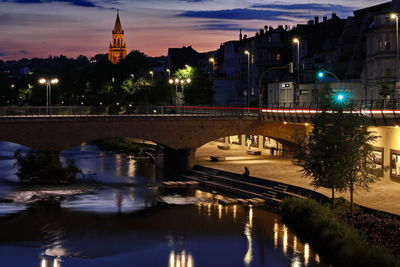 Illuminated bridge over river by buildings against sky at night