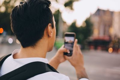 Rear view of young man photographing through smart phone in city