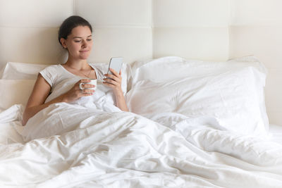 Beautiful young woman holding mobile phone and coffee cup while leaning on pillow in bed at home