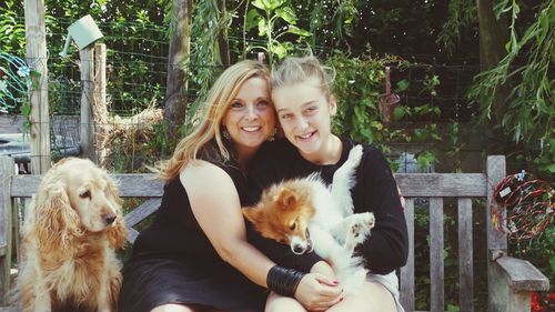 Portrait of smiling mother and daughter with dogs sitting on bench in park