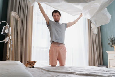 Young man and his funny dog are putting the bedding cover or mattress pad on the bed