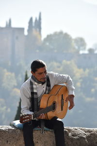 Mature man playing guitar while sitting on retaining wall