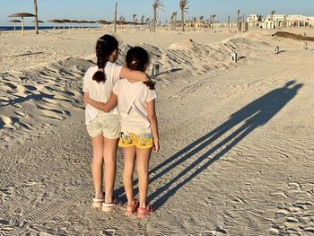 Rear view of two girls standing at beach with their shadow on the sand. 