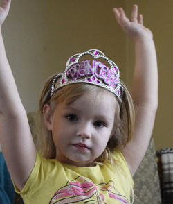 Close-up portrait of girl wearing crown with arms raised at home