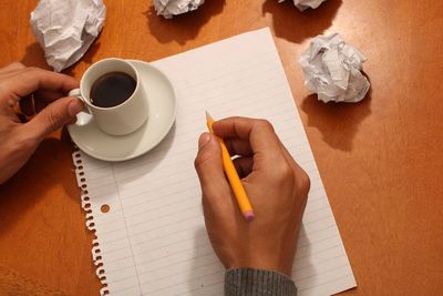 Cropped hand of person holding coffee while sitting with pencil and paper on desk