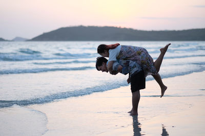 Side view of man piggybacking woman at beach against sky during sunset