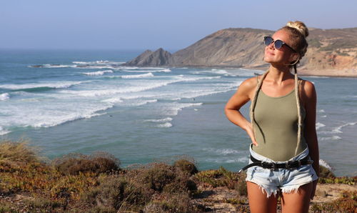 Young woman wearing sunglasses standing at beach against sky