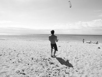 Rear view of shirtless man flying kite at beach against sky on sunny day