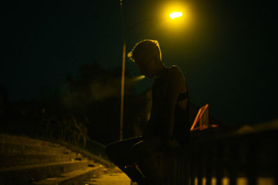 Silhouette woman smoking cigarette while sitting on railing against illuminated street light at night