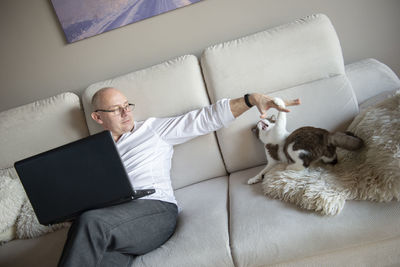 Mature man and cat using laptop on sofa at home during quarantine of coronavirus covid-19