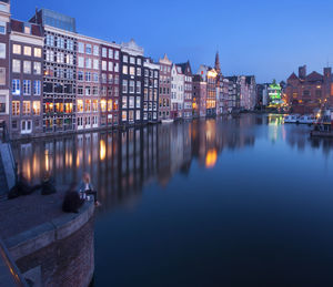 Illuminated buildings by river against sky at dusk