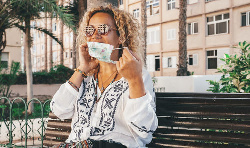 Smiling woman wearing mask while sitting bench outdoors